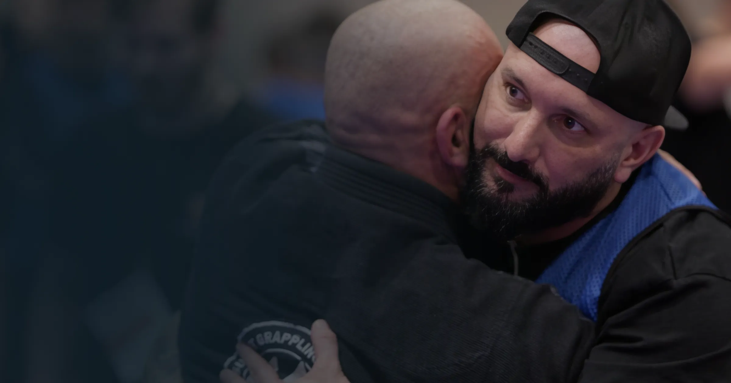 Two men embrace warmly, conveying camaraderie. One wears a dark cap and gi, the other is in a black gi with a patch. The background is softly blurred.